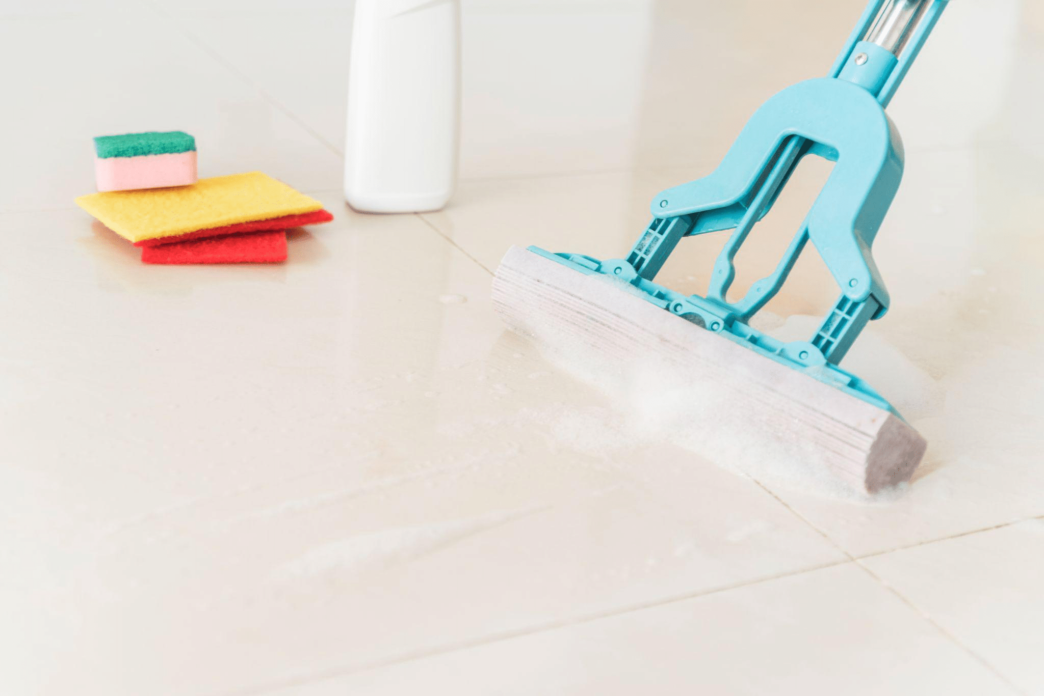 Blue mop cleaning a floor with soap suds, next to colorful cleaning sponges